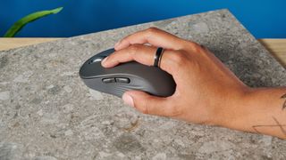 A black mouse with a scroll wheel, two side buttons, and embossed pads, with a logo, sits on a pink table with a blue background.