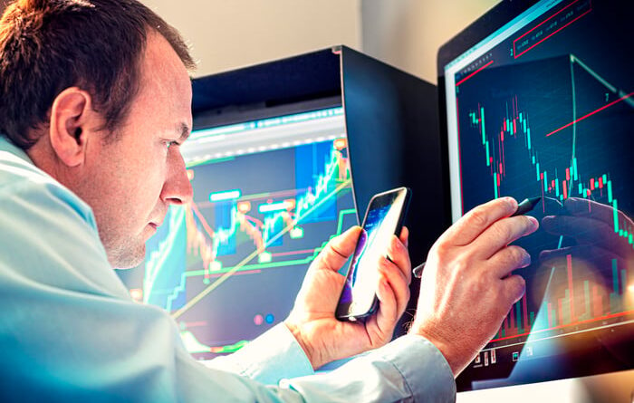 Professional money manager using pen and smartphone to analyze stock chart displayed on computer screen.