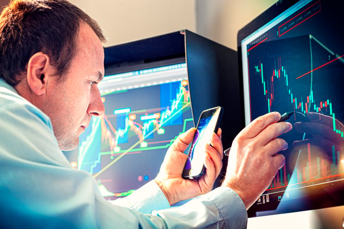 Professional money manager using pen and smartphone to analyze stock chart displayed on computer screen.